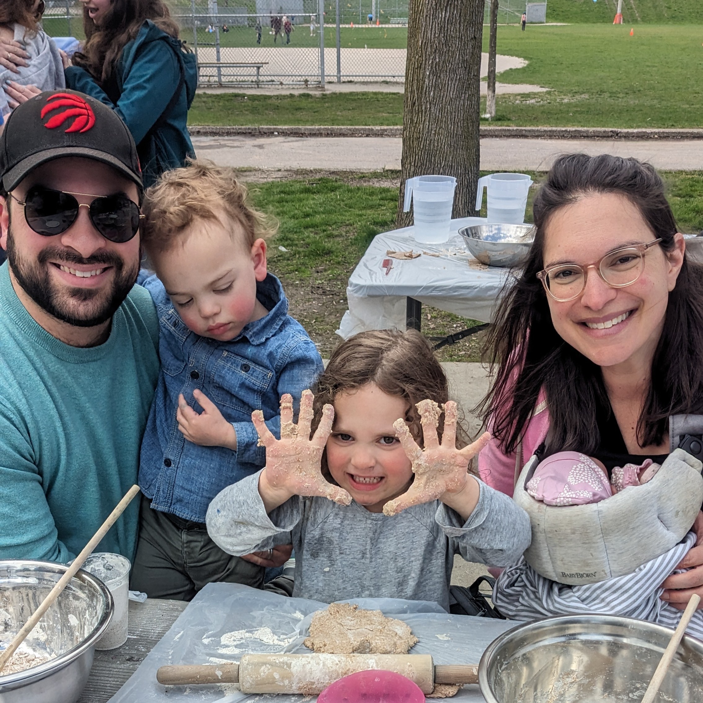 Smiling family at outdoor Matzah bake with young child showing off dough-covered hands